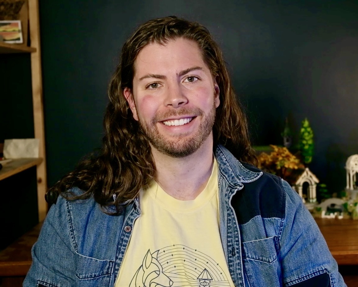 David Blocher, smiling, in a denim jacket, seated in front of a dark wall with shelves